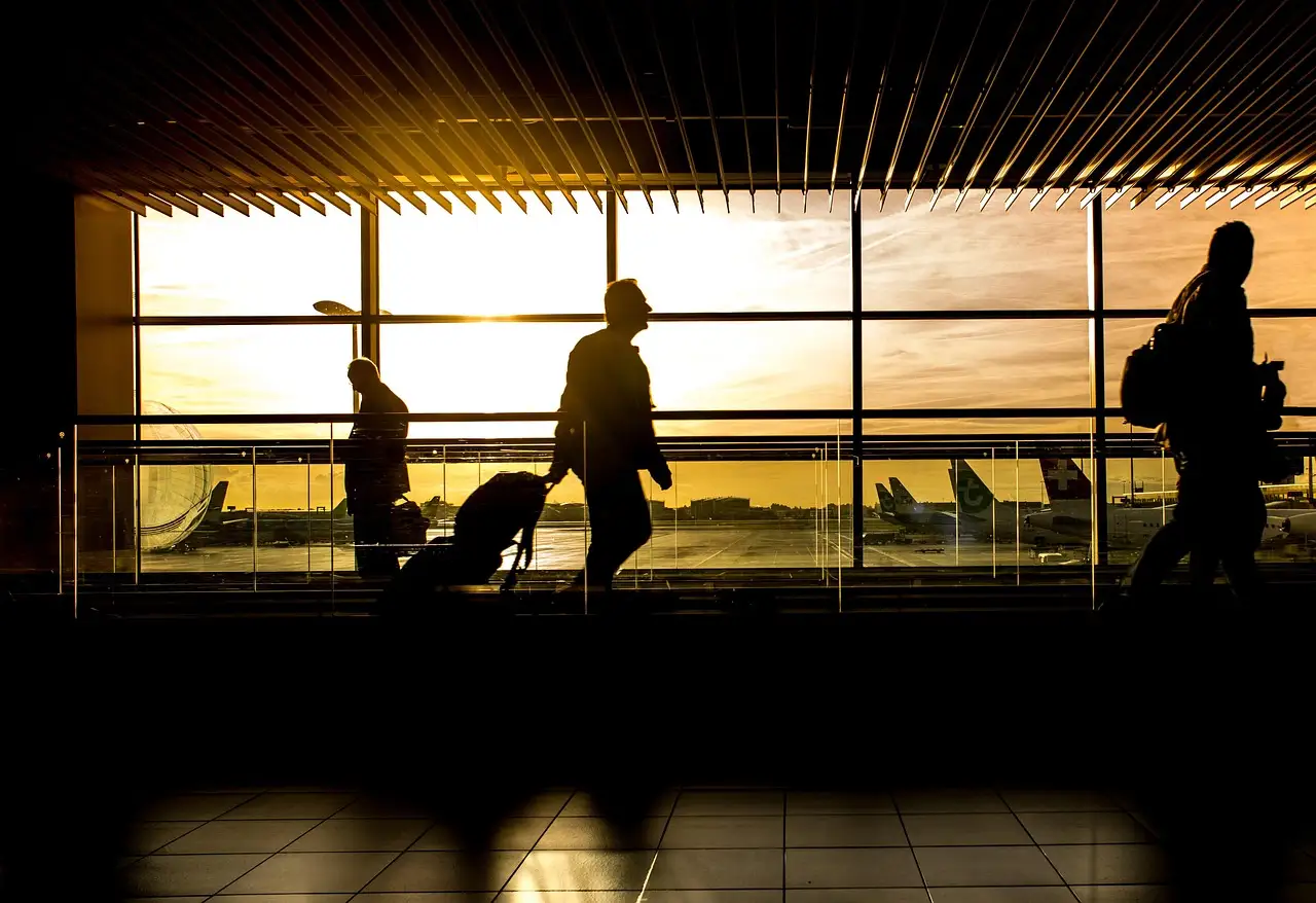 Silhouettes of travellers with luggage walking past large windows in an airport terminal at sunset