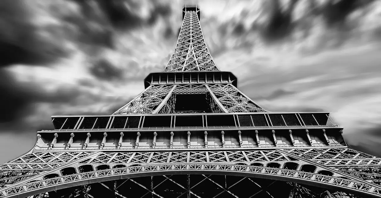 Black-and-white view up through the Eiffel Tower's iron lattice under a cloudy sky