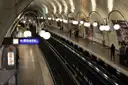 A curved, white-tiled Paris metro platform lined with globe lamps and an exit sign