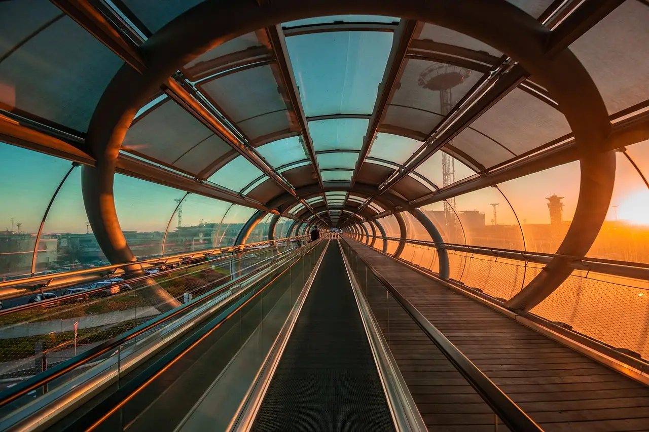 A glass-roofed airport walkway with a moving walkway lit by evening sun