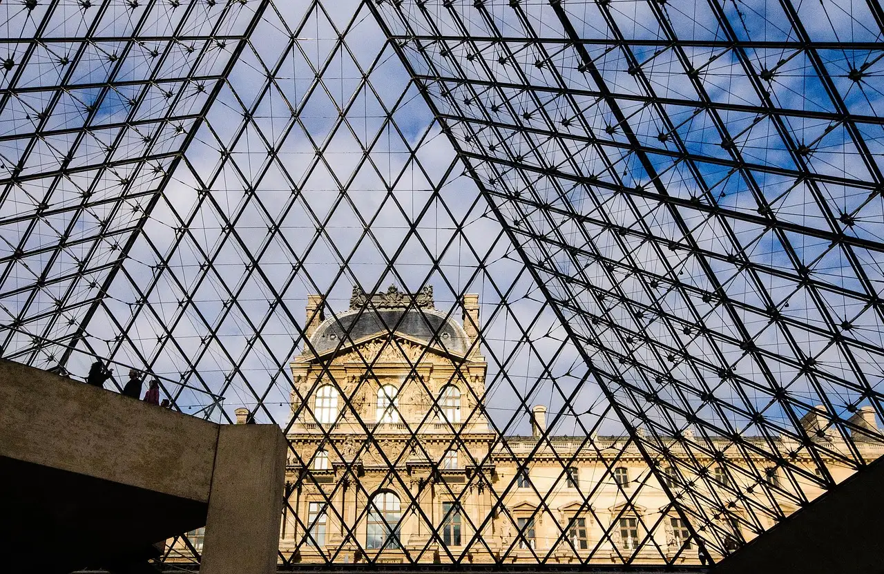 The Louvre's glass pyramid seen from below with the museum's stone wing behind it under a blue sky