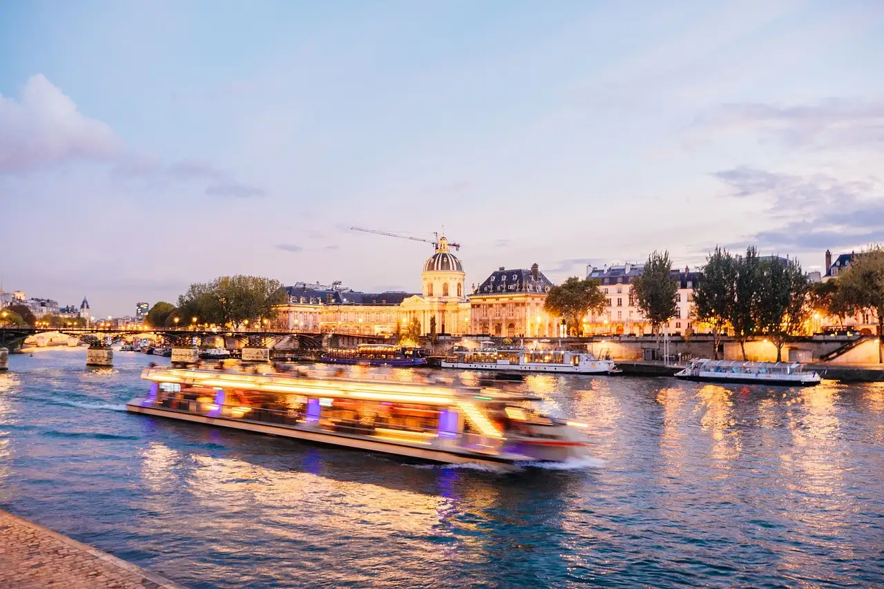 A lit-up river boat moving along the Seine at dusk, with the Institut de France behind